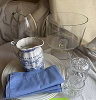 Photo of Jamie Oliver plates stacked at the bottom, thick glass vase and deep trifle dish behind, crystal-style blue and white creamer with floral design and chipped sugar bowl in front, blue napkin folded