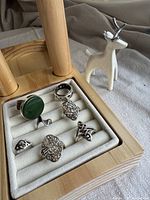 Six silver rings displayed on a wooden ring tray with white padding next to a white ceramic reindeer ring holder with silver antlers.