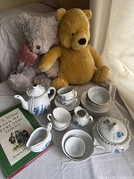 Two vintage teddy bears and child size tea time dishes with a Milne book on a cushioned surface near a window with curtain light.