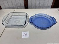 Two glass kitchen items on table: a clear glass square casserole dish with handles and floral pattern, and a blue glass round pie plate with handles.