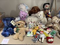 Image shows an assortment of teddy bears, Beanie Babies, and McDonald's Teenie Babies displayed on a table. Various colors and sizes of plush teddy bears along with multiple tagged Beanie Babies and plastic Teenie Baby figures are visible.
