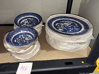 Stacked Churchill Willow blue and white plates and bowls in protective fabric covers on shelf.