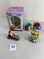 Lot items on table including Winnie the Pooh Anniversary Clock in box, Pooh snowglobe, two Mickey and Minnie mugs, and Grumpy figurine business card holder.