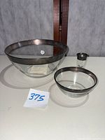 Set of one large crystal bowl with sterling silver rim, one small crystal bowl with sterling silver rim, and one shot glass with sterling silver rim on white surface.