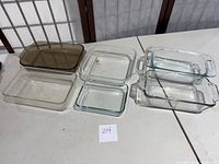Six glass casserole and refrigerator dishes arranged on a table showing different sizes and shapes including brown tinted and clear glass pieces.