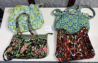 Four assorted quilted fabric bags with varied floral and geometric patterns displayed on a table with two brown leather chairs in background.