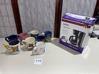 Eight assorted coffee mugs with various designs and colors along with the Rival 12 cup coffee maker box