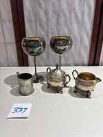 Full view of two engraved silver plated wine goblets, engraved MSC silver cup, Sheridan silver on copper creamer and sugar bowl on white surface.