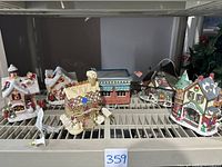 Six ceramic snow village and light-up houses on a metal shelf, including a gingerbread style house and several winter holiday themed buildings and figurines arranged close together.