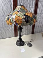 Tiffany style stained glass lamp on white surface showing full lamp with cord and foot pedal switch, leaf and berry pattern in orange, green, brown, and blue.
