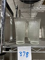 Two Jeanette Glass Finlandia tumblers shown side by side on a metal shelf, displaying the textured tree bark design and overall tapered shape with flared rim.
