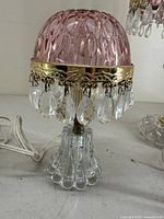Side and angle view of pink cranberry glass boudoir lamp with gold-tone metal trim and hanging crystal drops showing detailed pattern and clear glass base.