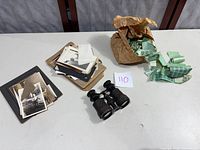 Photo showing a pile of assorted vintage black and white photographs, damaged binoculars, and a bundle of green stamps spilling from a brown paper bag.