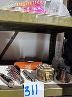 Photo showing a shelf with vintage pocket knives, a wooden box with nut crackers, a brass cricket cage, and a brass pitcher, also a belt buckle with decorative design.