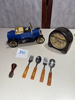 Photo showing the full lot including the blue vintage auto radio car model, four Bakelite children's utensils with decorative handles, a small wood handled tool, and the brown vintage shelf clock with scenic dial.