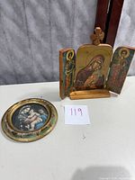 Photo of a hand-carved wooden triptych religious icon featuring central Virgin Mary with baby Jesus and two saints on side panels with a wooden cross on top. Also shown is a small framed circular religious artwork with an image of Madonna and Child with another figure in background, the frame is gilded with ornate detail.