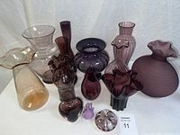 Photo showing collection of 13 art glass vases and vessels in amber, plum, purple shades with various shapes and sizes displayed on white fabric background.
