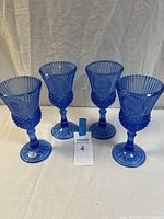 Front view of four cobalt blue glass stemmed goblets featuring embossed George Washington design, arranged in a row on a white surface with a background curtain.