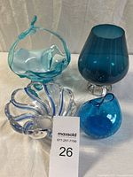 Photo showing four blue art glass pieces: aqua basket with handle, large blue pedestal vase, clear and cobalt swirl ribbed bowl, small cobalt crackle glass vase.