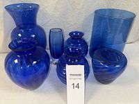 Collection of seven cobalt blue art glass vases including ribbed round vases, a tall cylindrical vase, and other smooth rounded shapes shown on a white backdrop.