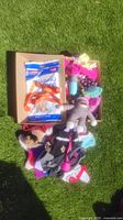 Box containing assorted dog clothing, a grey sock monkey plush toy, dog toys, orange leash, and training pads on grass.