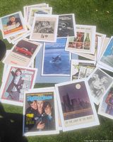 Wide shot showing collection of multiple vintage Rolling Stone magazine covers, vintage car and beverage advertisements, and an orca art print all unframed and mounted on foam core board displayed outdoors on grass