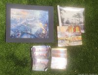 Overall assortment of Thomas Kinkade studio prints displayed on grass including large and small canvas prints and a box with small prints