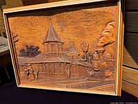 Frontal view of the wooden plaque showing a detailed engraving of an old steam locomotive and a station with figures waiting on the platform, taken with natural light highlighting the wood grain and carving detail.