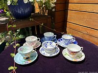 Top view of six vintage teacups with saucers arranged on dark surface, showcasing mixed floral and scenic designs with gold accents.