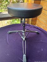 Front view of the adjustable silver drummer seat showing padded round black vinyl seat, adjustable metal support column and tripod base with rubber feet on a purple cloth.