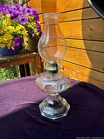 Clear antique glass oil lamp with metal burner and tall chimney on purple fabric in natural light, with flowers and wooden backdrop.