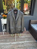 Front view of black leather jacket hanging on stand outdoors on wooden deck, showing jacket closed with button and flap pockets.