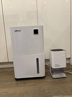 Front view of Britsou medium-sized white dehumidifier and smaller EasyAcc unit with 1200ML marking, both standing on floor.