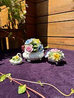 Wide shot of three delicate bone china flower figurines displayed on purple cloth against wooden background. Includes swan-shaped, round, and bowl-shaped porcelain pieces with multi-colored detailed flowers.