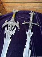 Top-down photo showing two decorative fantasy swords placed on a dark purple fabric surface for display.