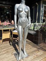 Front view of silver metallic full body female mannequin without head and arms, standing on rectangular metal base outdoors.