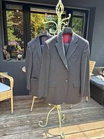 Two men's wool suits on hangers outdoors, one pinstripe and one solid gray, both with red lining inside.
