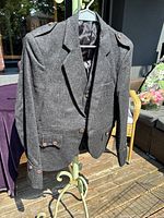 Grey wool kilt jacket hanging on a stand outdoors, showing front, lapels, sleeves with buttons, and shoulder epaulets.