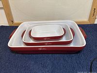 Three stacked Lagostina ceramic baking dishes in red with white interior, displayed on a blue cloth with a white background. The sizes range from small to large, visible with handles and logo.