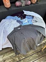 Photo showing assortment of men's dress shirts in various colors (grey, light blue, white) and patterns (plaid, check, solid) on hangers laid out on surface.