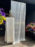 Photo of two sheer lace embroidered curtain panels hanging outdoors for display, showing delicate floral and leaf embroidery details and scalloped edges.