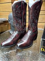 Front and angled view showing the red and black leather cowboy boots with decorative stitching and pointed toes on glass table, wood background.