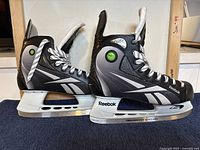 Side profile view of the black and silver Reebok ice hockey skates showing synthetic upper, white laces, and metal blades.