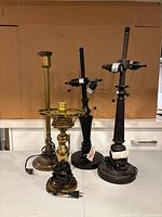 Four vintage lamp bases on a flat surface against cardboard background, including brass and matte black finishes, showing varied styles and coiled power cords.