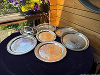 Seven silver plated serving trays arranged on a dark cloth surface outdoors, showing all trays including one with a lid. Trays have ornate engraved details and varying sizes.