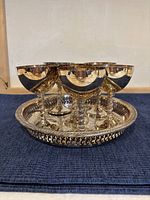 Photo showing entire set arranged with six goblets placed on a round silver plated tray with decorative etching, highlighting reflective surface and ornate edge details.