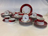 Full view of Stokes Plaid Christmas dinnerware set showing plates, cups, saucers, creamer, and sugar bowl all with festive red and white plaid patterns with gold accents.