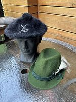 Image showing black angora Kangol cap on black mannequin head and green felt Bavarian hat on glass table against wood wall.