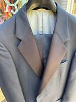 Close-up of the upper portion of the black tuxedo jacket showing the satin peaked lapels, chest pocket, and light beige lining inside the collar.