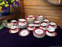 Full display of the dinnerware set arranged neatly on a dark cloth, showing cups, saucers, and plates with red and floral design and gold trim.